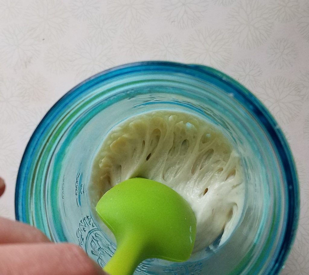 green spatula stirring small sourdough starter that is active in blue jar