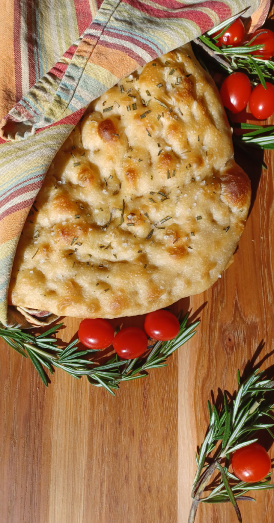 Sourdough focaccia loaf wrapped in striped towel surrounded by cherry tomatoes and rosemary.