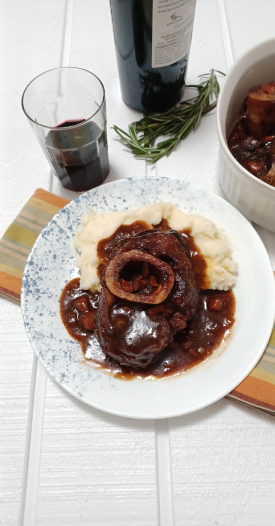 braised beef osso buco on plate with mashed potatoes sitting on striped towel and surrounded by wine and serving dish of the meal