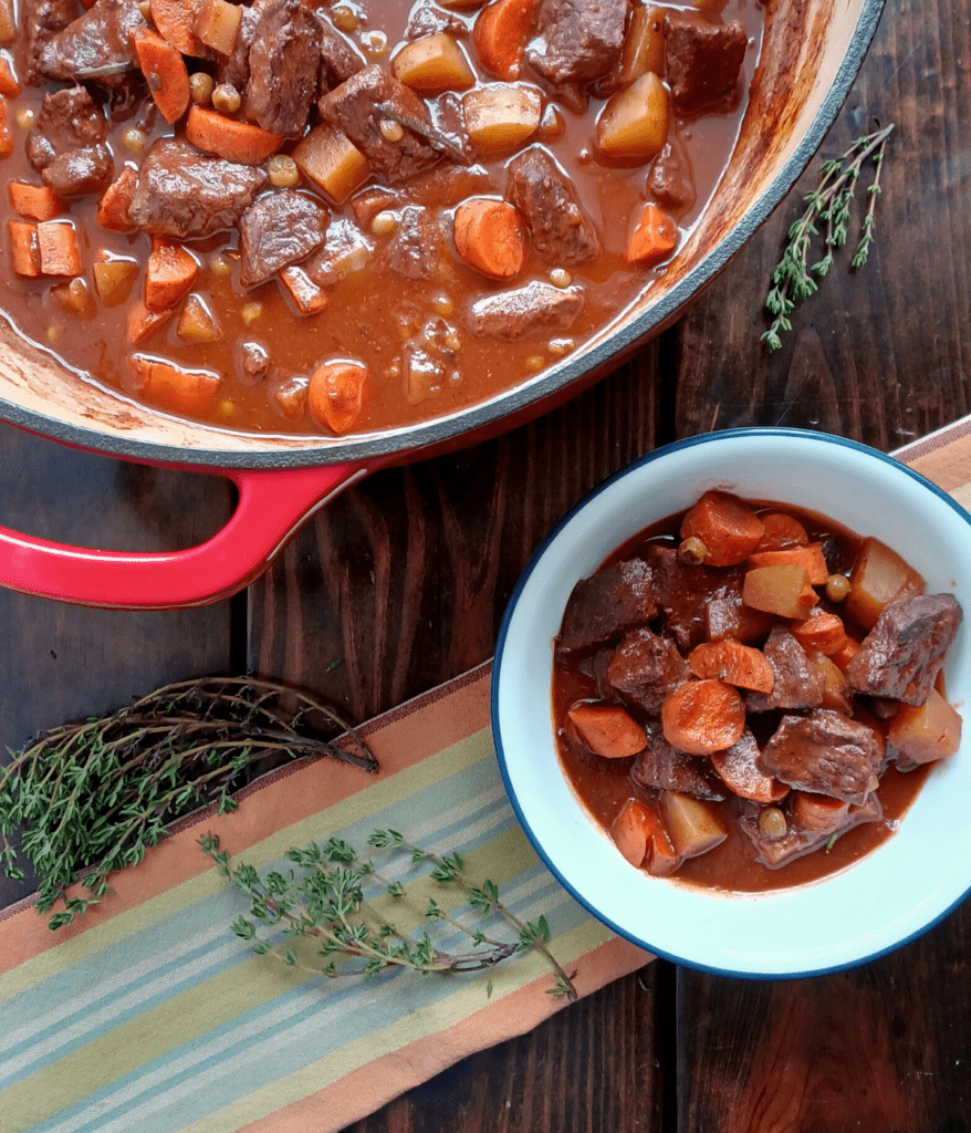 Homemade beef stew in bowl with blue rim on a striped towel with sprigs of thyme and in red dutch oven