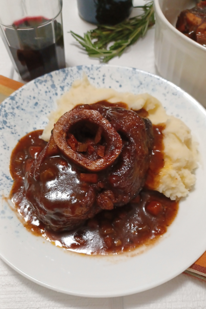 Braised beef osso buco on plate with mashed potatoes surrounded by glass and bottle of wine and rosemary