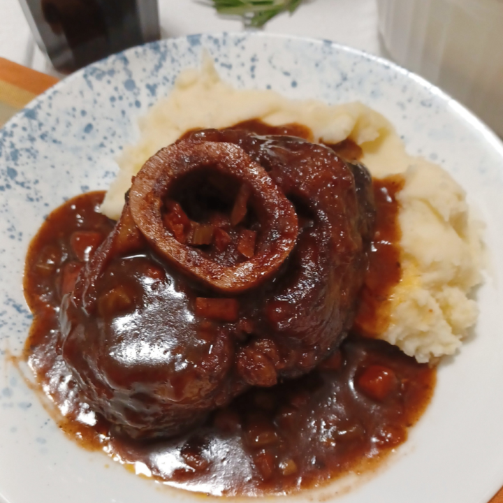 Braised beef osso buco on plate with mashed potatoes surrounded by glass and bottle of wine and rosemary