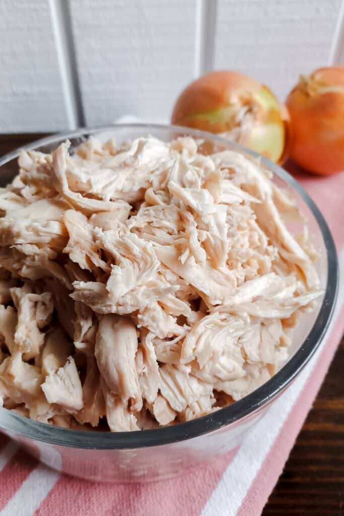 glass bowl of chicken that has been shredded on red striped towel with two yellow onions