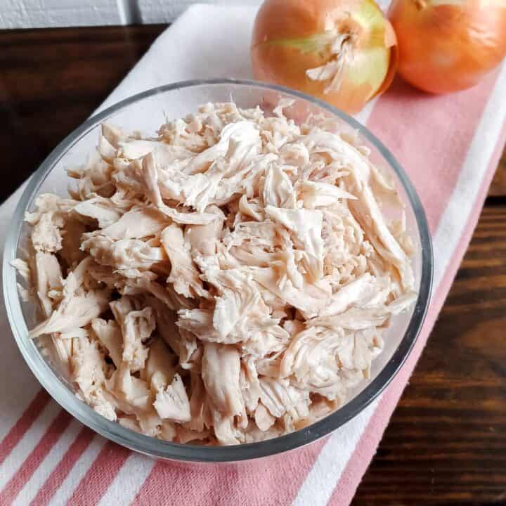 shredded chicken in a glass bowl on a red and white striped towel with 2 yellow onions