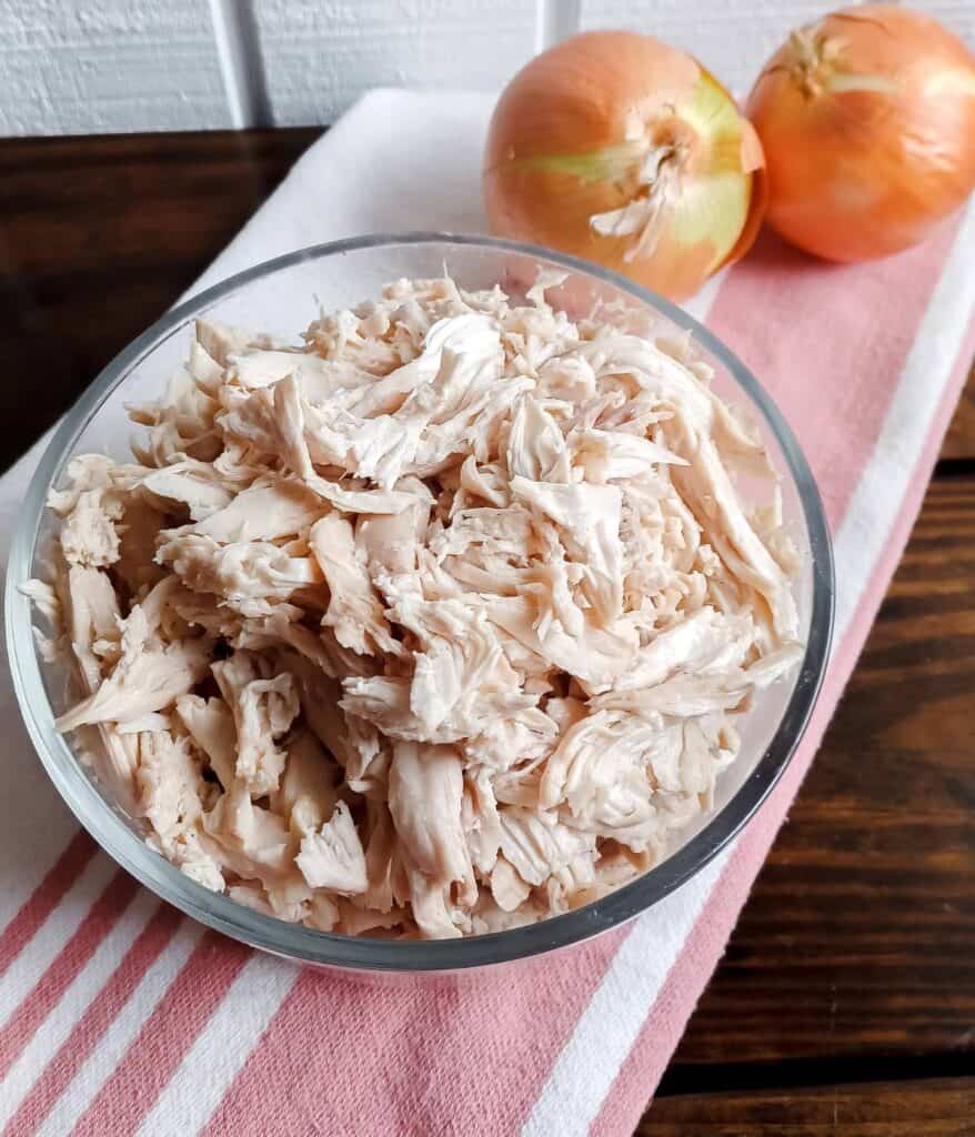 shredded chicken in a glass bowl on a red and white striped towel with 2 yellow onions