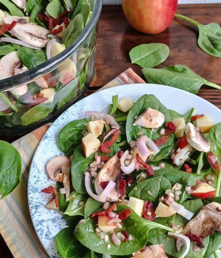 white plate with blue dots filled with spinach, apple, bacon, mushroom, and shallot dressing in a maple-Dijon vinaigrette in front of a glass bowl with same ingredients and loose spinach leaves and an apple surrounding both.