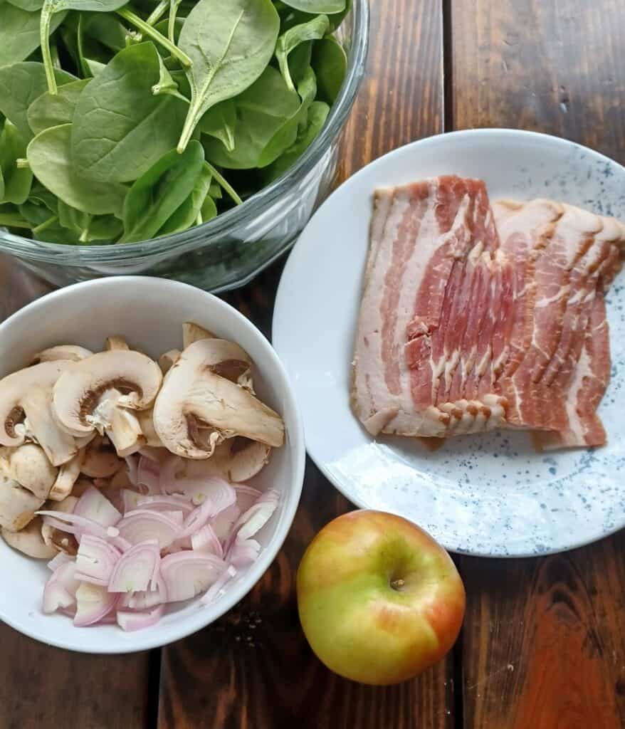 spinach in glass bowl, sliced mushrooms and shallots in a white bowl, uncooked bacon on a white plate and a whole apple