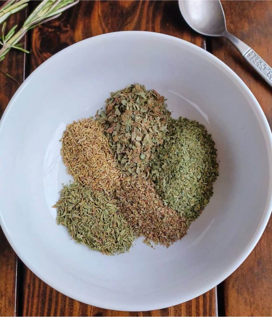 Dried, basil, oregano, thyme, rosemary, and marjoram in a white bowl on wooden table with spoon and rosemary springs