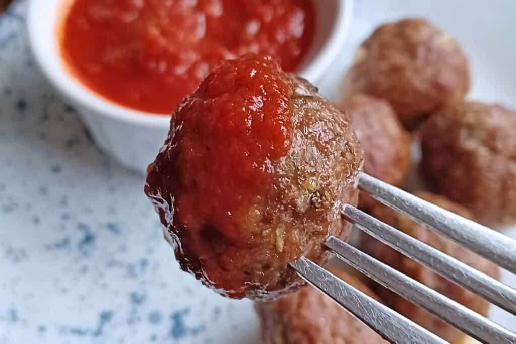 Baked homemade meatball on a fork with tomato sauce on it. bowl of tomato sauce in background with other meatballs