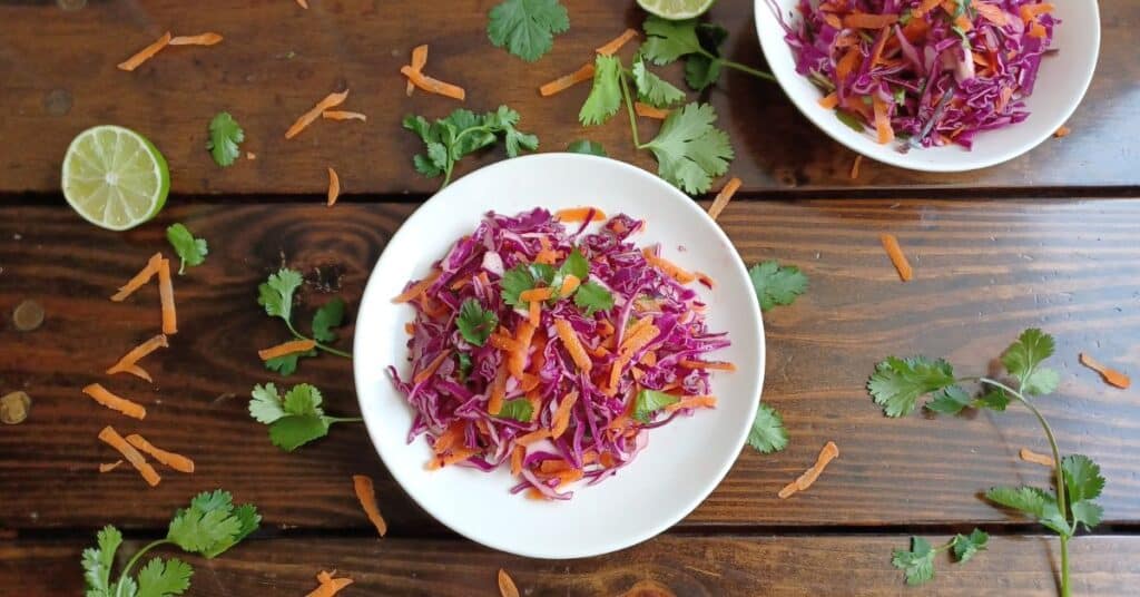 Two white bowls filled with cilantro lime slaw surrounded by a lime half, shredded carrots, and cilantro leaves