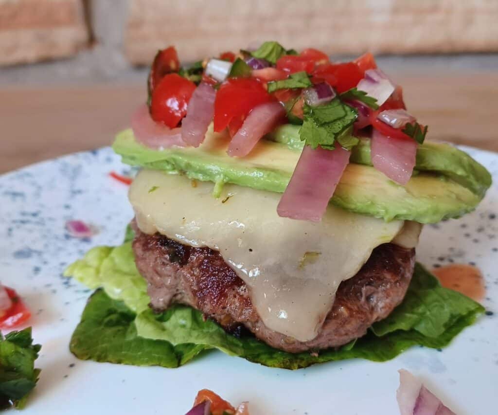Salsa burger topped with cheese, avocado, pickled red onions, and pico de gallo on white plate with blue dots