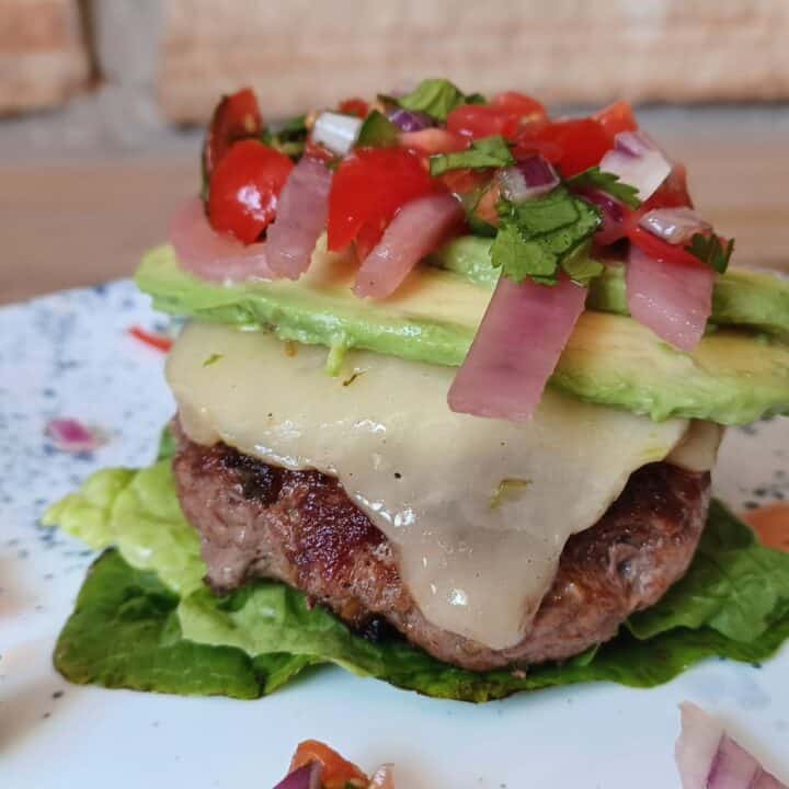 Salsa burger topped with cheese, avocado, pickled red onions, and pico de gallo on white plate with blue dots